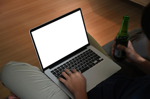 Cropped shot of man holding bottle of beer and using laptop on sofa