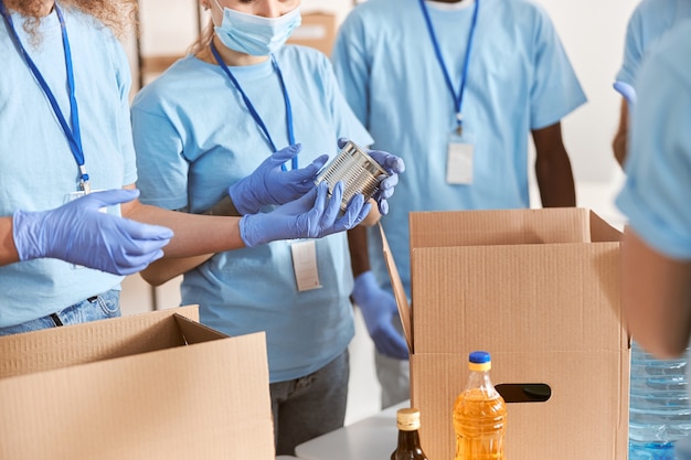 Cropped shot of volunteers in blue uniform protective masks and gloves sorting packing canned food