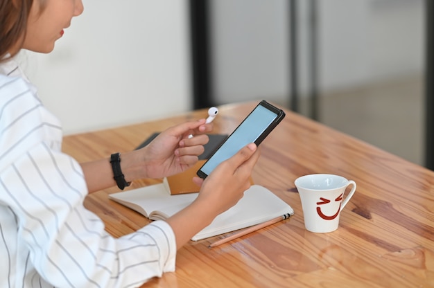 Cropped shot of a woman in striped shirt holding white blank screen smartphone and wireless earphone in hand sitting at the wooden desk.