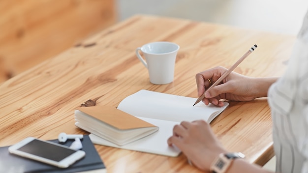 Cropped shot of young beautiful woman in striped shirt writing/taking note sitting at the wooden working desk.