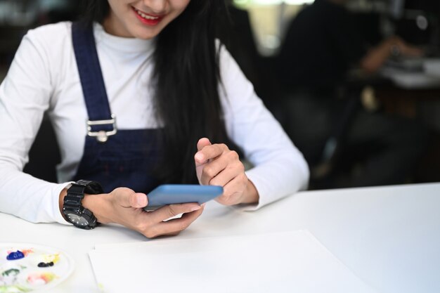 Cropped shot of young happy female artist or designer using smart phone at her workstation.