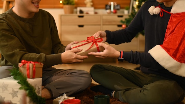 Cropped shot of young man giving Christmas present to his friend while sitting in cozy living room