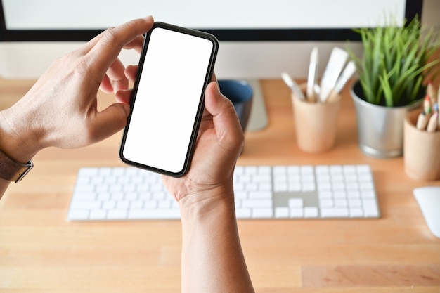 Cropped shot of young man holding blank screen mobile phone on desk workspace.