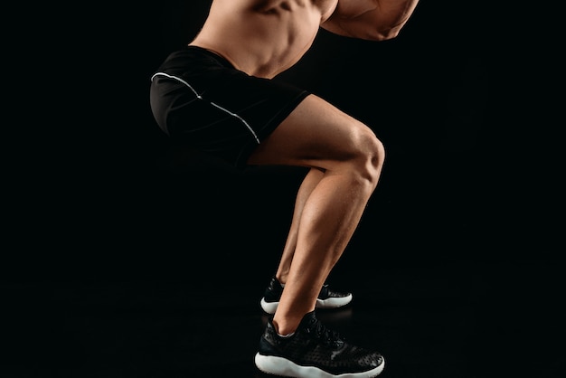 Cropped view of bodybuilder with muscular legs squatting isolated on black