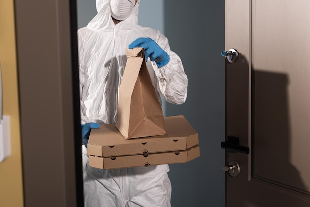 Cropped view of delivery man in hazmat suit and latex gloves holding package and pizza boxes near