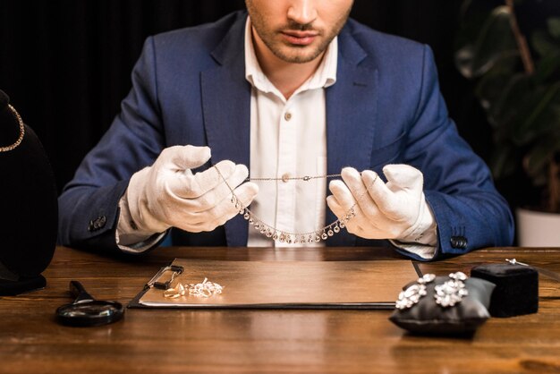 Cropped view of jewelry appraiser holding necklace near jewelry on board on table in workshop