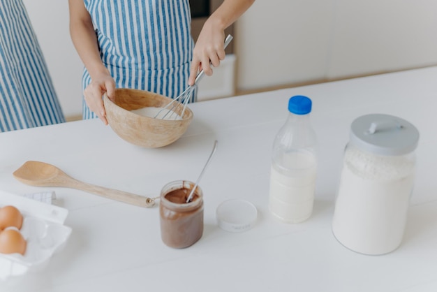 Cropped view of kid in apron whisks ingredients in bowl with beater busy cooking and helping mother to prepare cake Dough milk eggs wooden spatula and chocolate on table