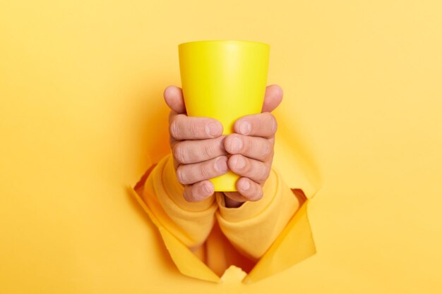 Cropped view of man holding coffee to in mug in both hands through hole in paper wall isolated on yellow background