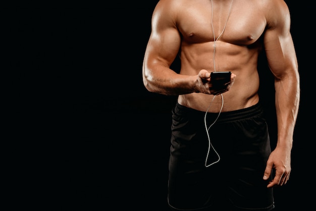 Cropped view of sexy bodybuilder with muscular torso listening music with earphones on smartphone isolated on black