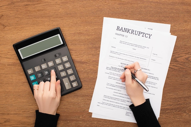 Cropped view of woman filling in bankruptcy form and using calculator on wooden background