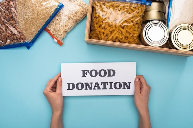 Cropped view of woman holding card with food donation lettering near cans and groats in zipper bags