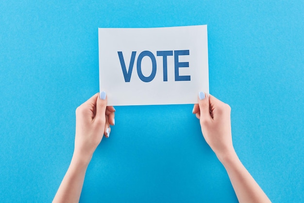 Cropped view of woman holding white card with vote lettering on blue background