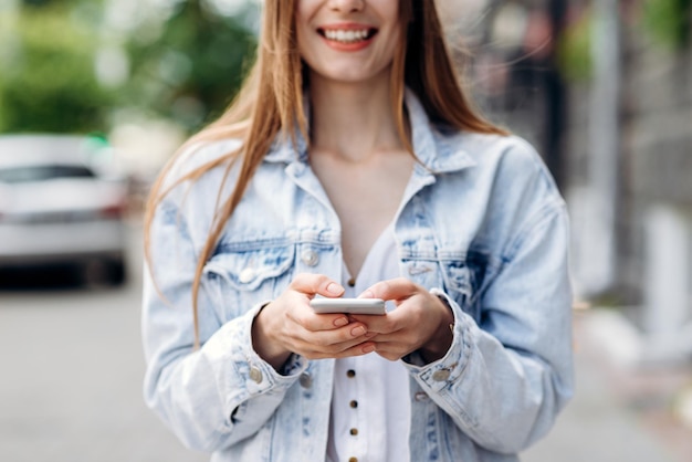 Cropped view of the woman wearing jeans jacket texting on the smart phone while walking in the street in a sunny day