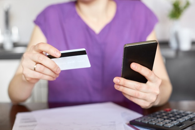 Cropped view of young woman in purple clothes sits at table, surrounded with documents, calculator, holds smart phone and plastic card, checks her bank account online, works at home. Payment concept