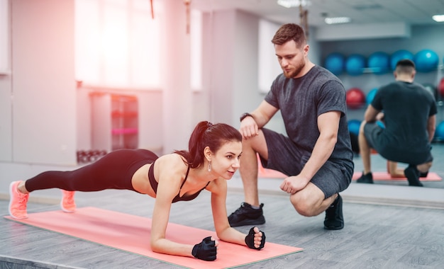 Crossfit female doing push-ups exercises with elbow forearms on a pink mat in the gym. Dark-haired fitness woman with trainer doing plank, elbow strap. Close-up