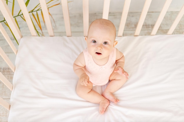 A crying baby girl in a blanket in a crib on a white cotton bed of six months has woken up and is calling for her mother