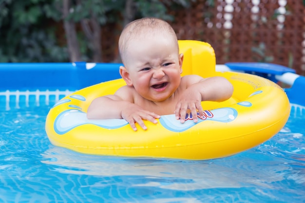 A crying newborn baby of 10-12 months swims in a yellow swimming ring. Frame swimming pool in the backyard of the house