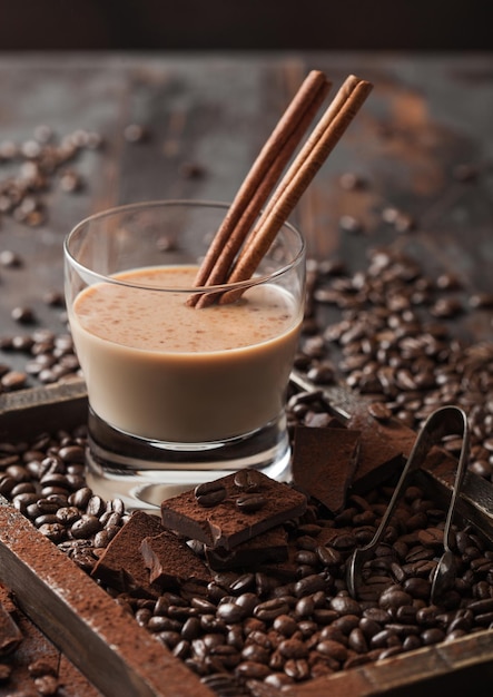 Crystal glass of Irish cream baileys liqueur with cinnamon coffee beans and powder with dark chocolate in wooden tray on dark wood background Top view