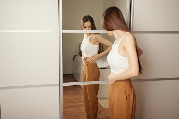 The cult of a beautiful body public opinion imposed stereotypes A teenage girl is looking for fat on her body stands at home in front of a mirror