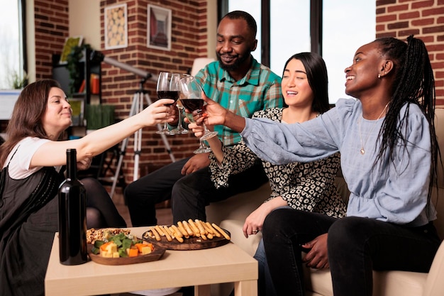 Culturally diverse group of adults, men, women toasting, seated at living room couches, drinking glasses of wine. Ethnically different people bunch enjoying alcohol, eating bread sticks.