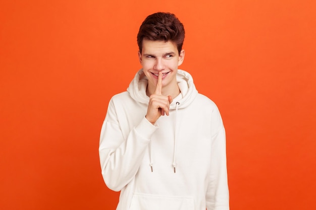 Photo cunning young man in casual style hoodie showing silence gesture, holding finger near lips and smiling, keep calm knowing secrets. indoor studio shot isolated on orange background