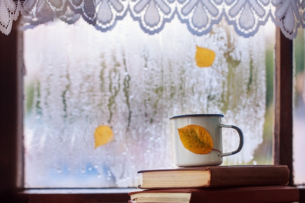 Cup of autumn tea or coffee, and yellow leaves on rainy window