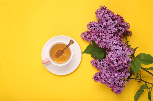 Cup of black aromatic tea with lemon and branches of blooming lilacs on yellow background Top view Flat lay