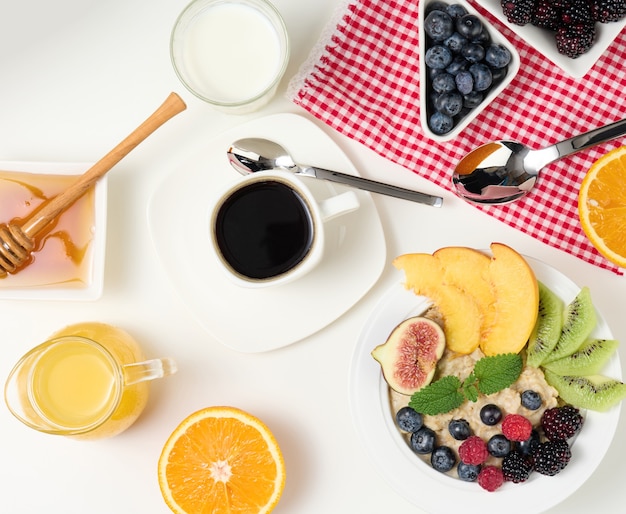 Cup of black coffee, a plate of oatmeal and fruit, honey and a glass of milk on a white table, healthy morning breakfast, top view