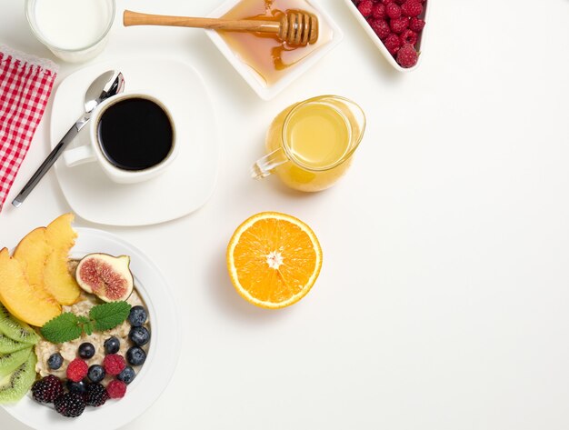 Cup of black coffee, a plate of oatmeal and fruit, honey and a glass of milk on a white table, a healthy morning breakfast