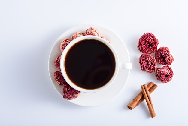 Cup of black coffee with roses on plate, cinnamon sticks on white table