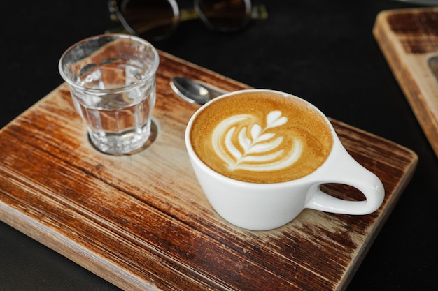 Cup of cappuccino with latte art in cozy cafe coffee and glass of water on wooden board