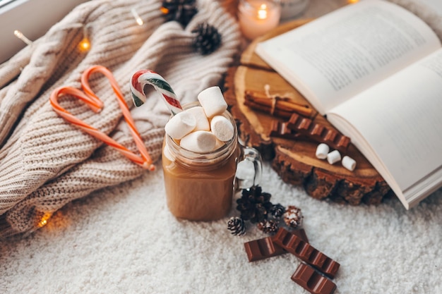 A cup of cocoa with marshmallows chocolate an open book lamp lights in a cozy homely atmosphere on the windowsill Christmas aesthetics