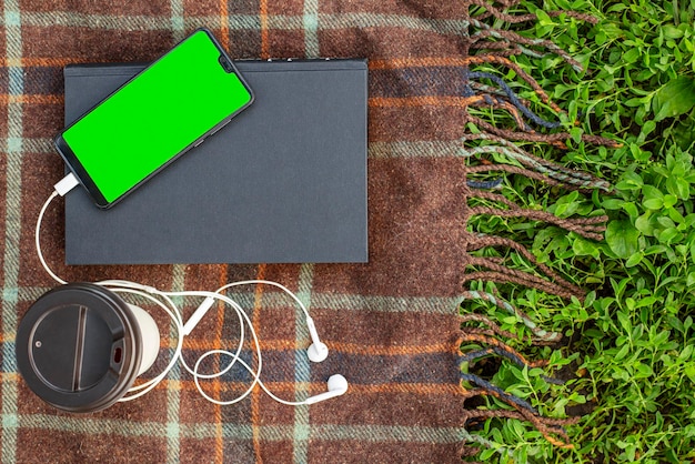 Cup of coffee book and smartphone with headphones on blanket in park or garden