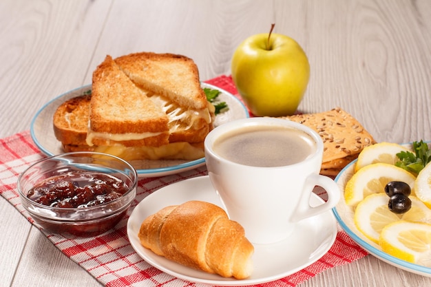 Cup of coffee and croissant on saucer, glass bowl with strawberry jam, toasted slices of bread with cheese, apple, cookies with grains of sunflower seeds, cut lemon on plate and red napkin. Top view