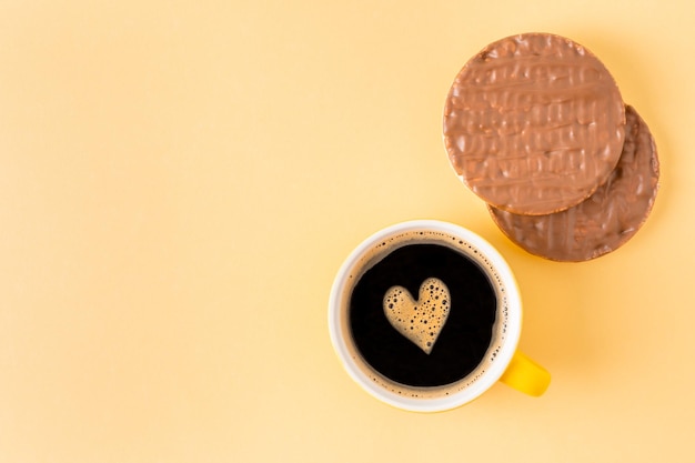 Cup of coffee decorated with foam heart near chocolate covered rice cakes on yellow background, top view, empty space