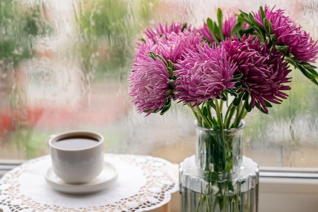 A cup of coffee and flowers in the vase standing next to a rainy window.