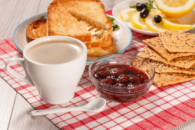 Cup of coffee, glass bowl with strawberry jam, toasted slices of bread with cheese, lemon and cookie. Good food for breakfast.