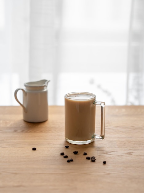 A cup of coffee laltte with milk on a wooden table with coffee beans and milk jug against the background of a window