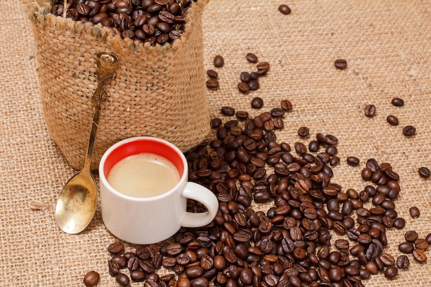 Cup of coffee, a metal spoon and roasted coffee beans in a canvas sack on sackcloth. Top view.