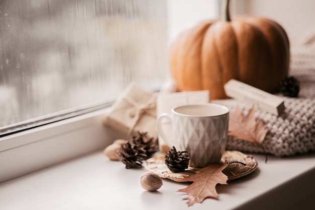 Cup of coffee, pumpkin, dried autumn leaves on window.