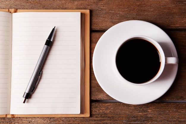 Cup of coffee on saucer with notebook and pen on wooden table background