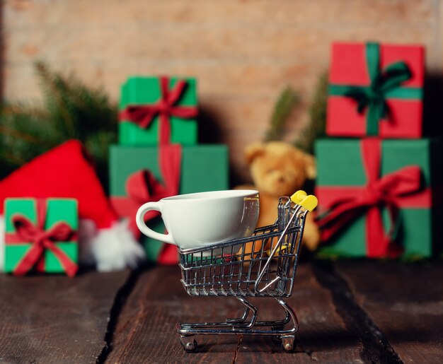 Cup of coffee and shopping cart next to Christmas gifts on wooden table