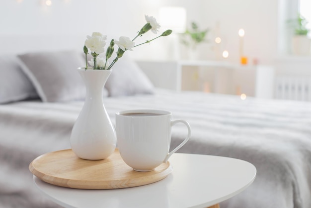 Cup of coffee and white flowers in vase on table in bedroom