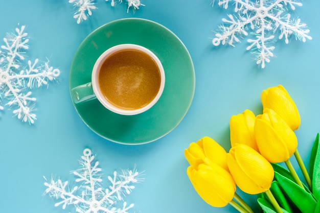 Cup of coffee with artificial yellow tulips and snowflake ornaments on blue background