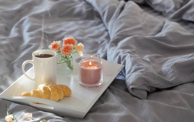 Cup of coffee with croissant  and pink roses on bed in  bedroom