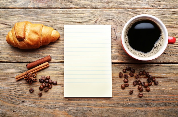 Cup of coffee with fresh croissant and blank sheet of paper on wooden table top view