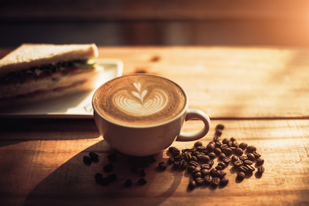 A cup of coffee with heart pattern in a white cup and Sandwich on wooden table background 