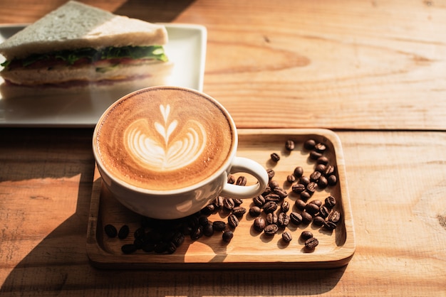 A cup of coffee with heart pattern in a white cup and Sandwich on wooden table background