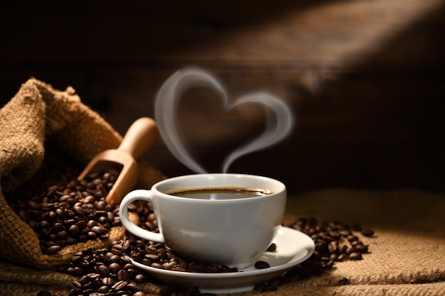 Cup of coffee with heart shape smoke and coffee beans on burlap sack on old wooden background