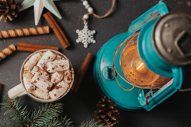 A cup of coffee with marshmallows, Christmas decor and a kerosene lamp on dark gray 